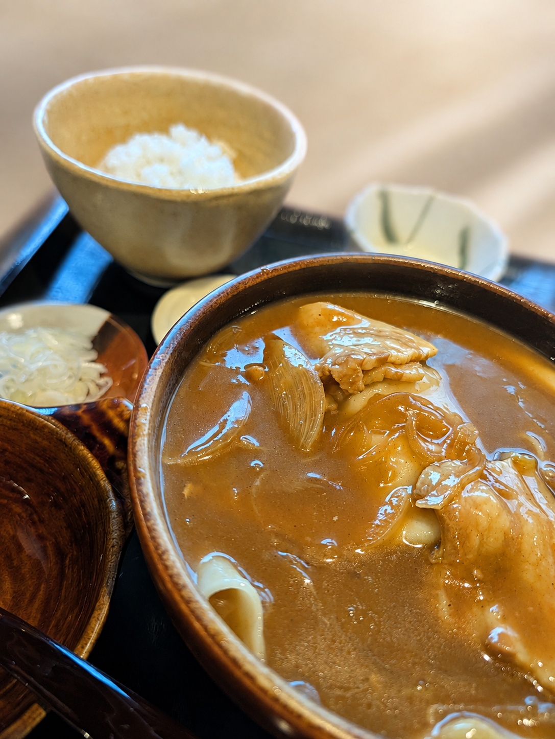 木の器に入ったカレーのひもかわうどん。奥にご飯の茶碗と漬物の小皿