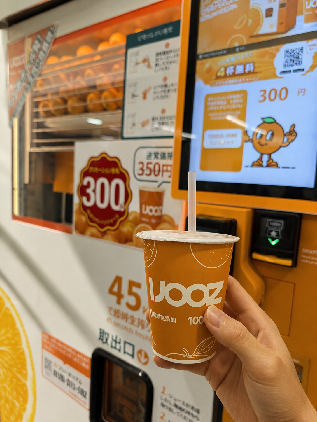 IJOOZの自販機の前で、生搾りオレンジジュースのカップを持っている