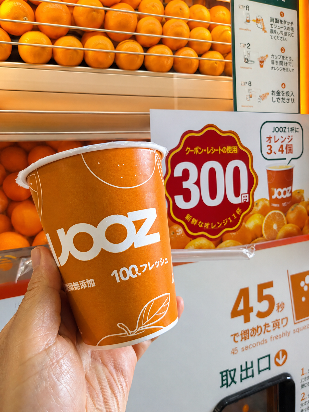 IJOOZの自販機の前で、生搾りオレンジジュースのカップを持っている