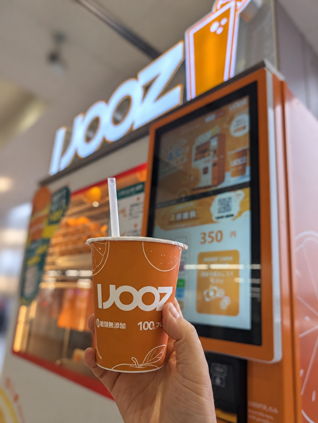IJOOZの自販機の前で、オレンジジュースのカップを持っている