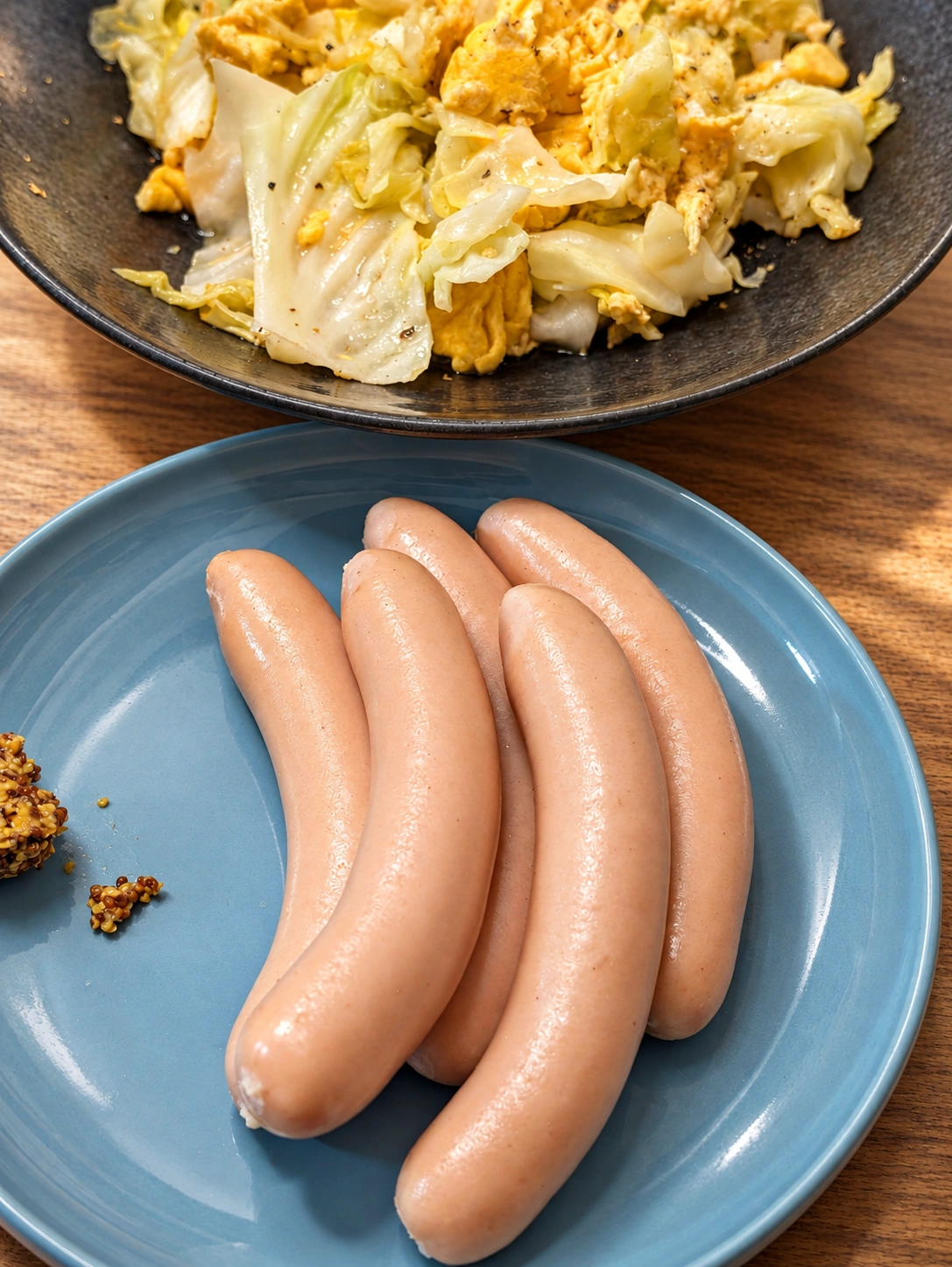 青い皿にスワンレイクビールのソーセージ4本と粒マスタード、奥にキャベツと卵の料理（卓上・光などをAIで加工）